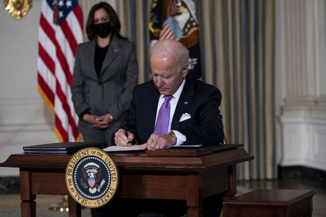 Biden sits at a desk and holds a pen to sign an order
